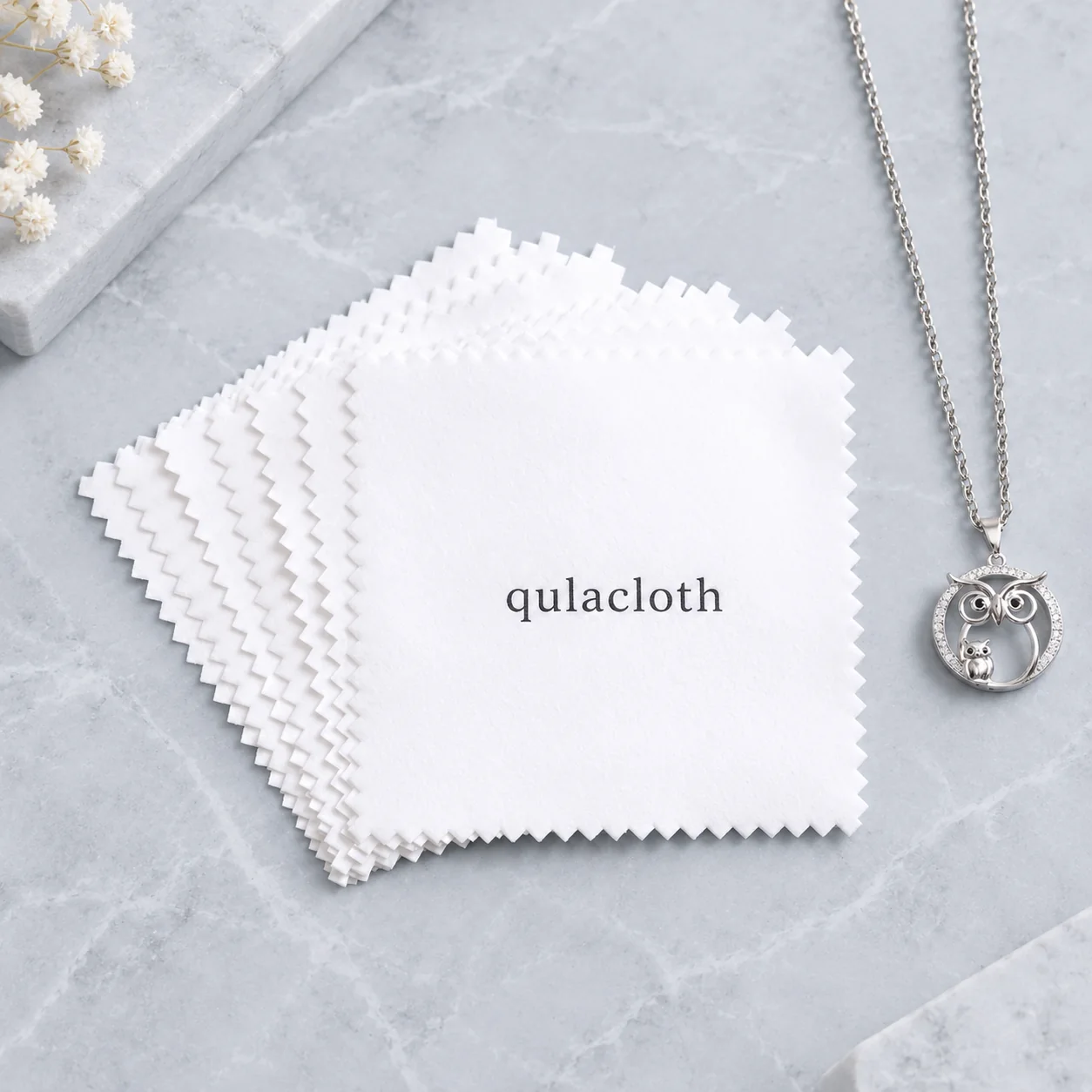 Stack of white 8 x 8 cm zigzag-edge jewelry cleaning cloths with black Qulacloth logo and silver owl necklace
