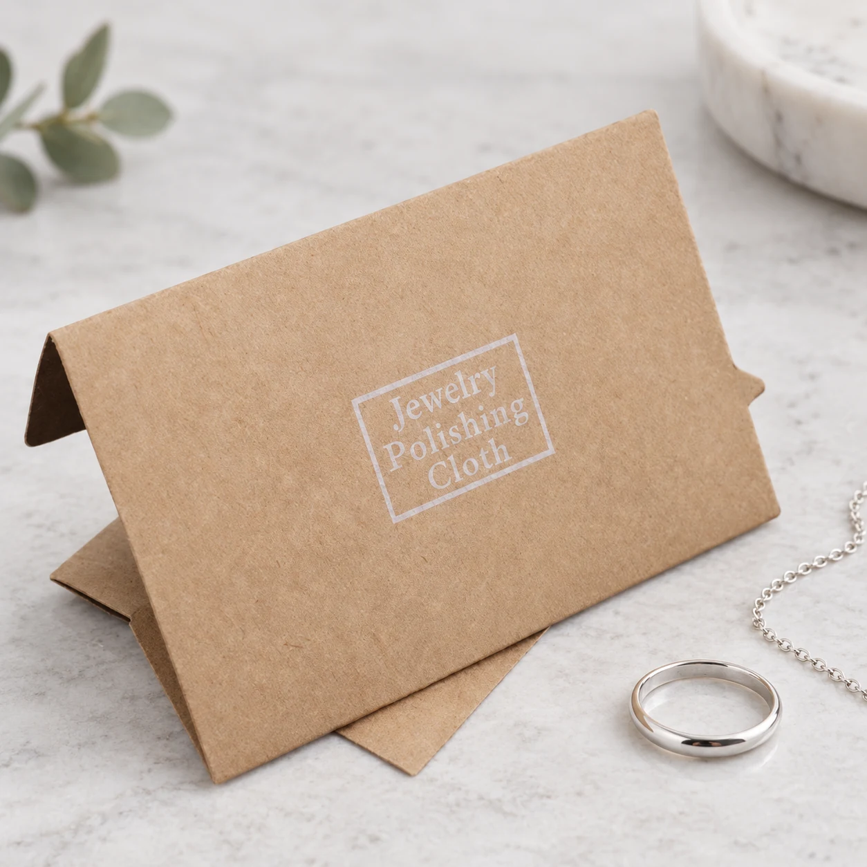 Kraft paper jewelry polishing cloth envelope beside a silver ring and chain for wholesale jewelry care kits