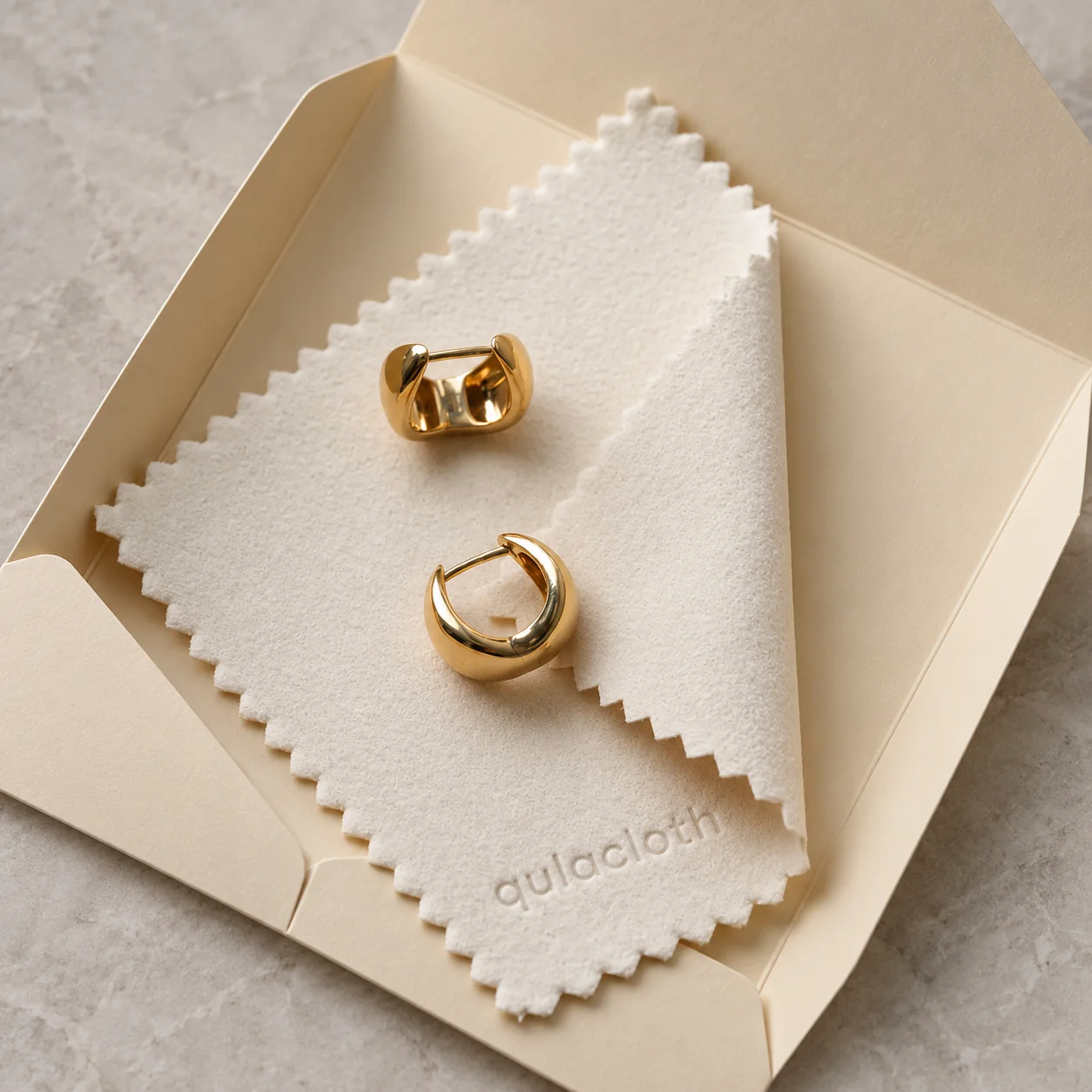 White zigzag-edge jewelry polishing cloth with Qulacloth debossed logo and gold hoop earrings